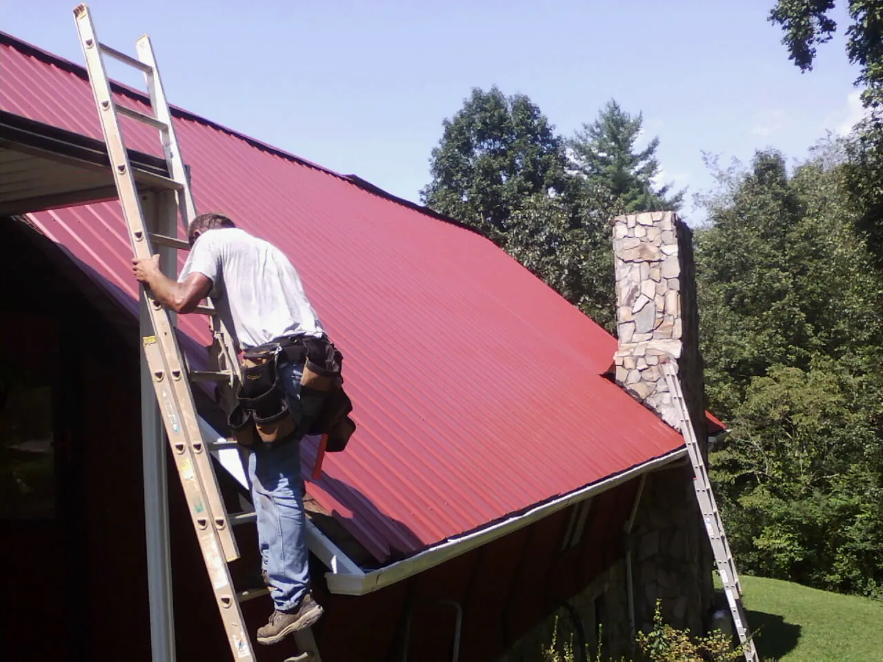 Roofer on a ladder beside a red metal roof and stone chimney — Soffit & Fascia Repair by Roofing Brothers in Vauxhall